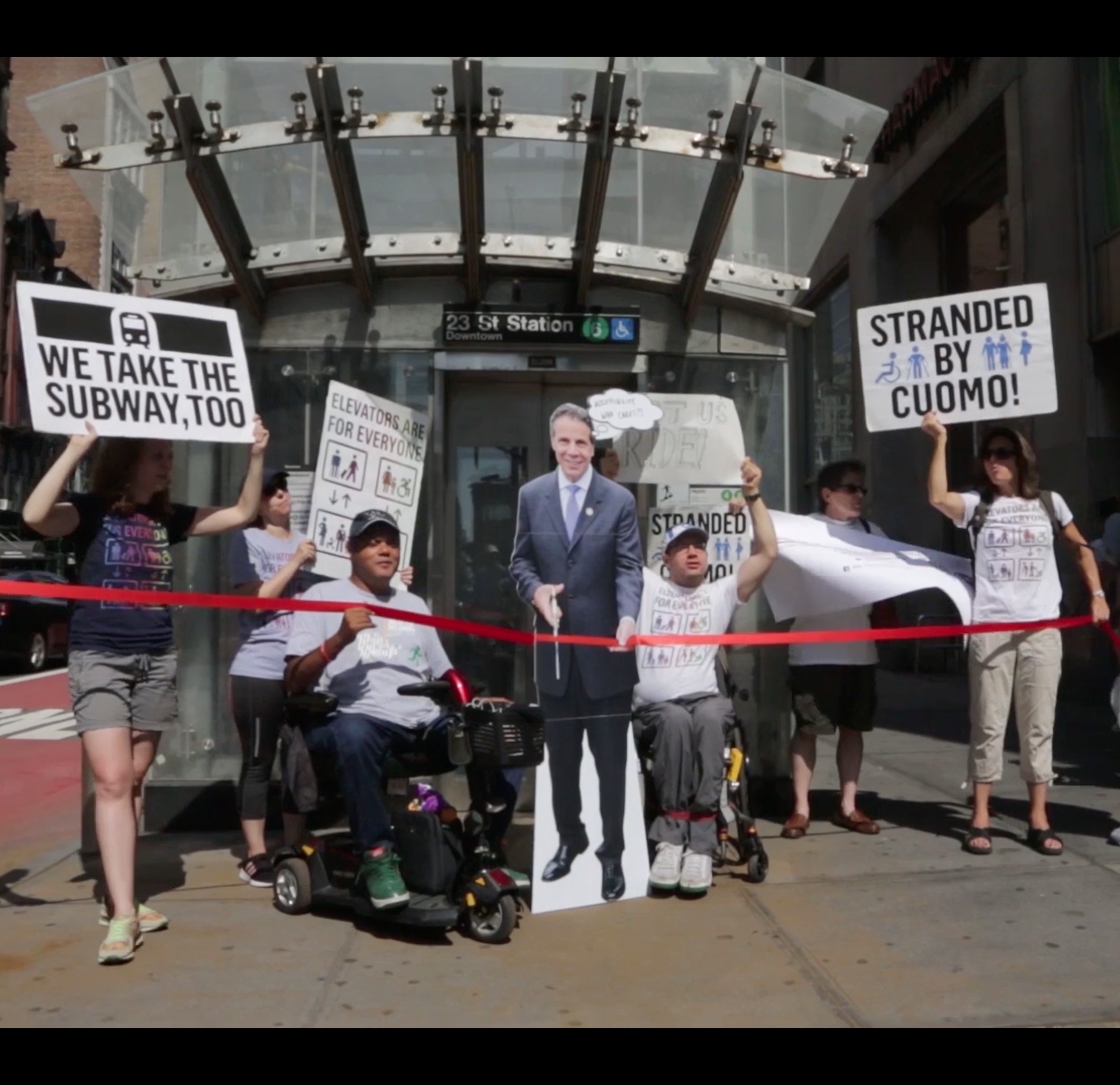 activists carry posters that read "stranded by cuomo", "We take the subway too" and "elevators are for everyone" next to a cardboard cutout of governor Cuomo cutting a ribbon
